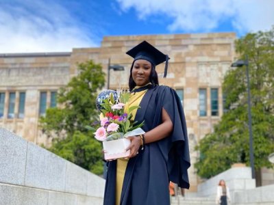 A young African woman stands in graduation cap and gown and holding flowers outside UQ's sandstone Great Court.